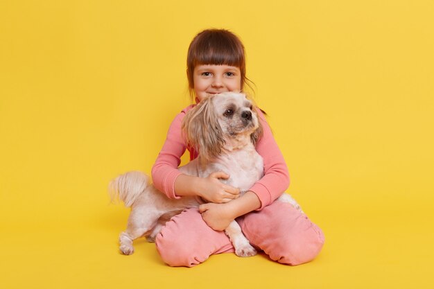 Linda niña abrazando a su perro mascota aislado en amarillo