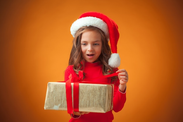 La linda niña alegre con gorro de Papá Noel y regalo sobre fondo naranja