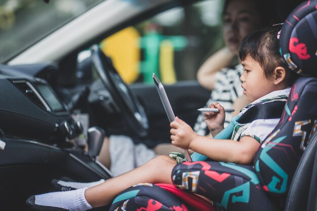 Linda niña asiática sentada en un auto con su mamá y viendo dibujos animados