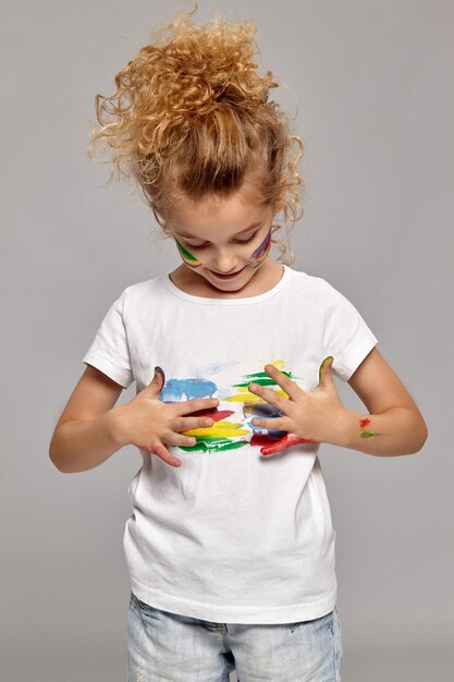 Linda niña con un cepillo en su hermoso corte de pelo, vestida con una camiseta blanca. Ella está manchando su camiseta, sobre un fondo gris.