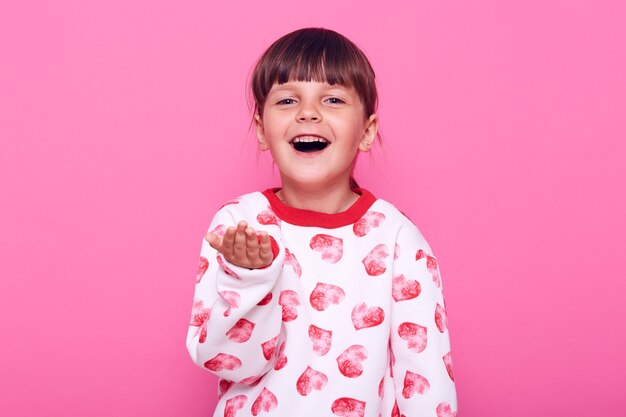 Linda niña en edad preescolar feliz con sudadera casual con corazones y palma de la mano, con expresión satisfecha, buen humor, aislada sobre pared rosa.