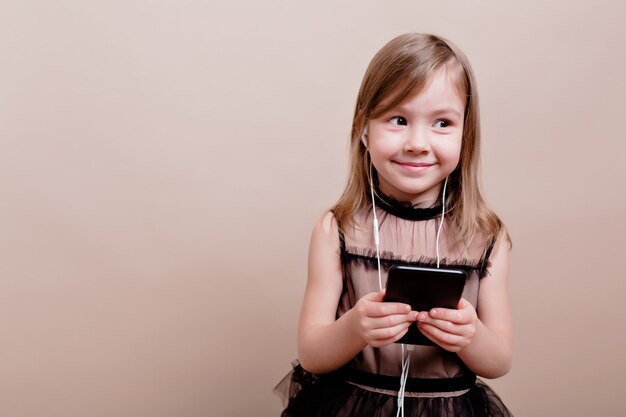 Linda niña encantadora con una sonrisa encantadora escuchando música en auriculares y posando en una pared aislada con grandes emociones, lugar para el texto