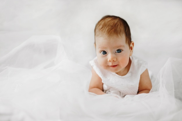 Linda niña está acostada sobre el vientre con grandes ojos azules abiertos, vestida con traje blanco