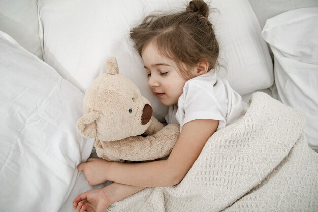 Una linda niña está durmiendo en una cama con un osito de peluche.