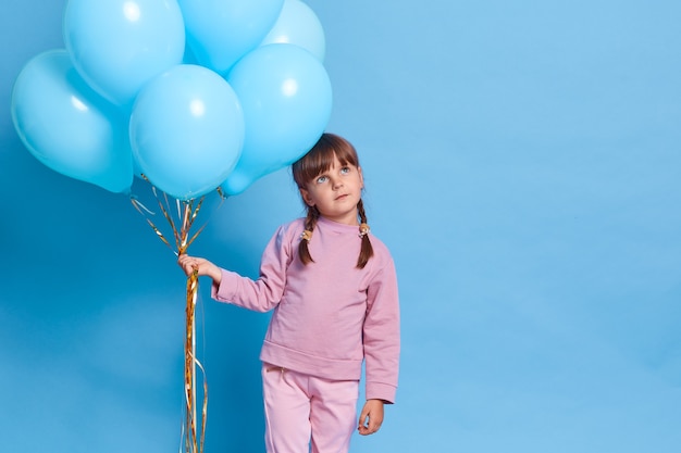 Linda niña europea con cierre de rosa, niño con coletas mirando con expresión facial pensativa, sueña con algo agradable, sosteniendo un montón de globos de helio, contra la pared azul.