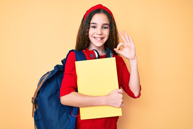 Linda niña hispana con mochila de estudiante y auriculares sosteniendo un libro haciendo el signo de ok con los dedos sonriendo amigablemente gesticulando excelente símbolo