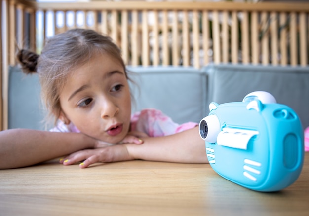 Una linda niña mira la cámara de juguete azul de sus hijos para imprimir fotos al instante.