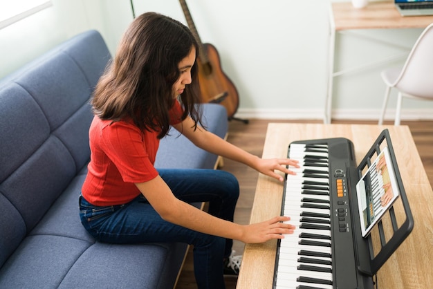 Linda niña preadolescente siguiendo las instrucciones de su profesor de música y aprendiendo a tocar el piano. Chica hispana tomando lecciones de arte en una videollamada en línea