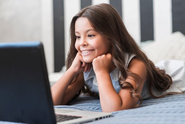 Linda niña sonriente acostada en la cama viendo el video en la computadora portátil