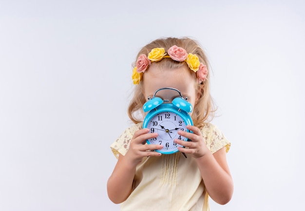 Una linda niña vestida con camisa amarilla en diadema floral que muestra el tiempo mientras sostiene el despertador azul sobre una pared blanca