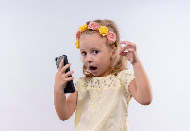 Una linda niña vestida con camisa amarilla en diadema floral sorprendente mientras sostiene el teléfono móvil en una pared blanca