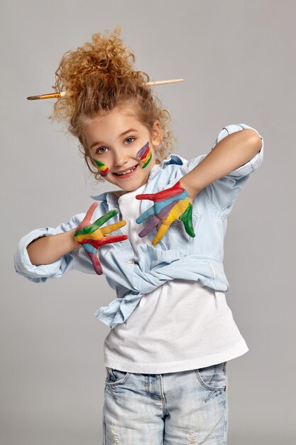 Una linda niñita con un cepillo en su elegante corte de pelo, vestida con una camisa azul y una camiseta blanca. Ella está posando con los brazos y las mejillas pintados, mirando a la cámara y sonriendo, sobre un fondo gris.