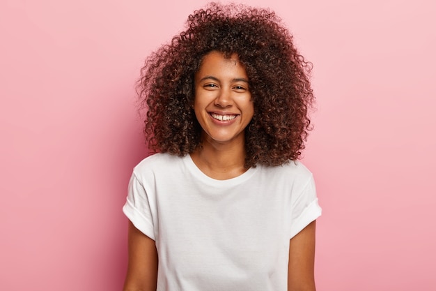 Linda novia feliz sonríe con alegría, tiene dientes blancos y uniformes, se ve, disfruta de un buen rato con su amante, escucha bromas divertidas, usa camiseta blanca, modelos contra la pared rosada. Concepto de etnia