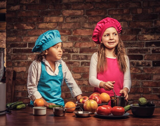 Linda pareja de cocineros. Un niño pequeño con cabello castaño rizado vestido con un uniforme de cocinero azul y una hermosa niña vestida con un uniforme de cocinero rosa cocinando juntos en una cocina contra una pared de ladrillos.