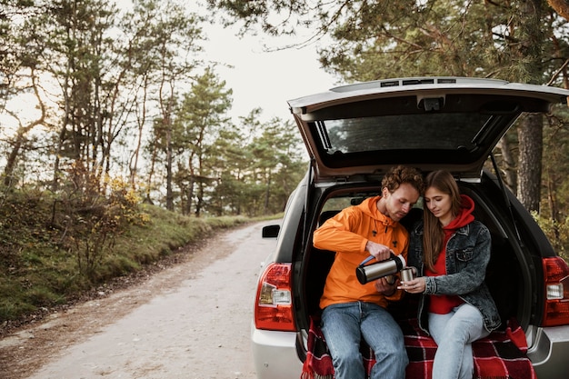 Linda pareja disfrutando de bebidas calientes en el maletero del coche