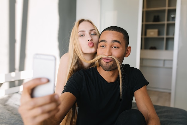 Linda pareja haciendo selfie en teléfono en la cama en apartamento moderno. Chico guapo divirtiéndose con el pelo largo y rubio de su novia, enviando un beso, alegría, estado de ánimo alegre