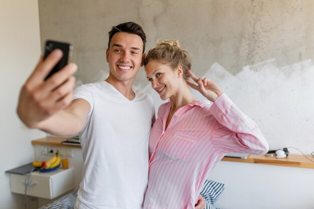 Linda pareja joven divirtiéndose en el dormitorio por la mañana, hombre y mujer haciendo foto selfie en pijama