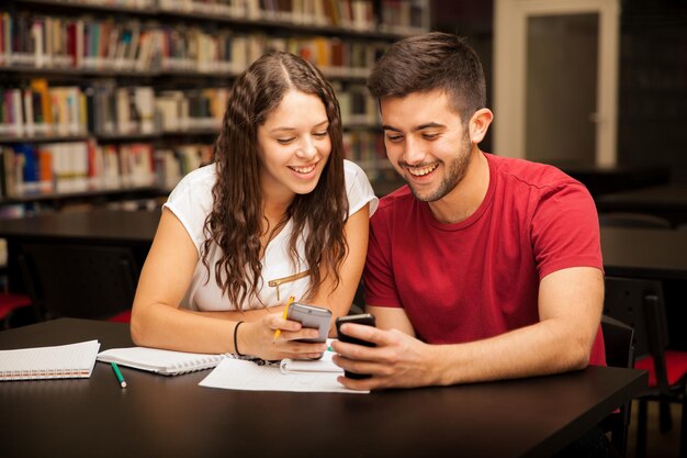 Linda pareja joven de estudiantes universitarios usando sus teléfonos inteligentes y estudiando en la biblioteca