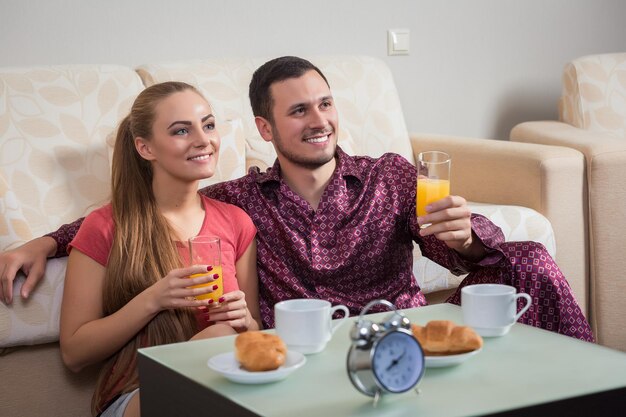 Linda pareja joven relajándose en casa en el suelo al lado del sofá desayunando, comiendo croissants y bebiendo jugo de naranja.