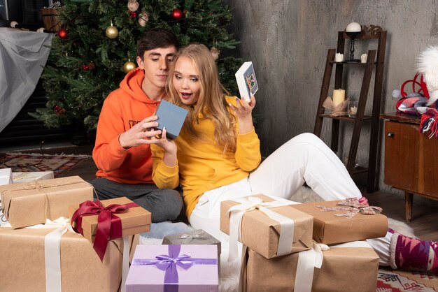 Linda pareja joven sentada y abriendo regalos cerca del árbol de navidad