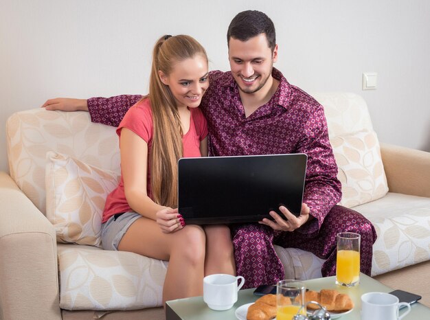 Linda pareja joven en el sofá desayunando, comiendo croissants, bebiendo jugo de naranja frente a una computadora portátil