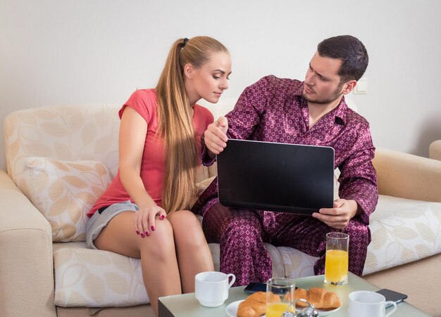 Linda pareja joven en el sofá desayunando, comiendo croissants, bebiendo jugo de naranja frente a una computadora portátil