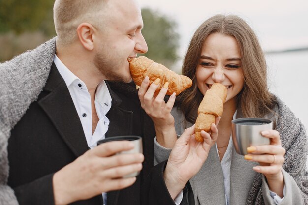 Linda pareja en un parque. Dama con un abrigo gris. Personas con termo y croissant.