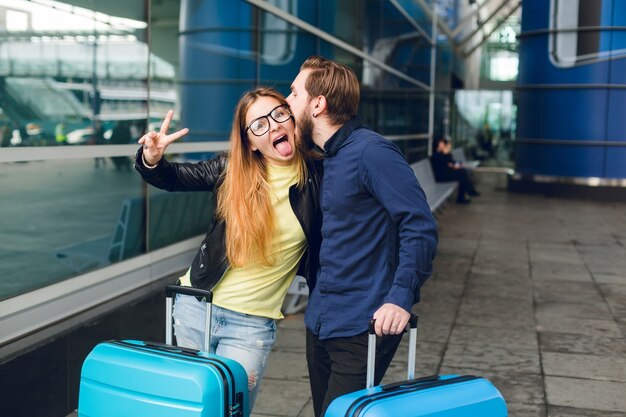 Linda pareja con sutcases está de pie afuera en el aeropuerto. Tiene cabello largo, lentes, suéter amarillo, chaqueta. Viste camisa negra, barba. Se abrazan y simulan juntos.