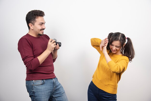 Linda pareja tomando fotografías con la cámara y posando sobre una pared blanca.