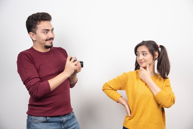 Linda pareja tomando fotografías con la cámara y posando sobre una pared blanca.