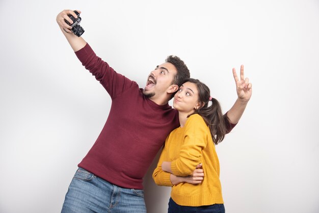 Linda pareja tomando selfie con cámara y posando sobre una pared blanca.