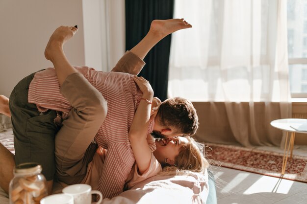 Una linda pareja vestida con ropa de color pastel está acostada en la cama y de buen humor está jugando en apartamentos luminosos.