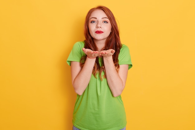 Foto gratuita linda romántica mujer pelirroja de pie y enviando amoroso beso de aire a la cámara, demostrando afecto, vistiendo una camiseta verde