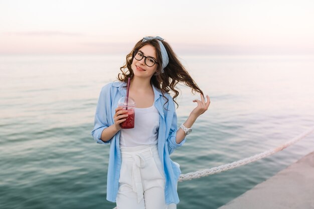 Linda señorita en vestido blanco y chaqueta azul posando juguetonamente en el muelle del océano y beber refrescos en la mañana