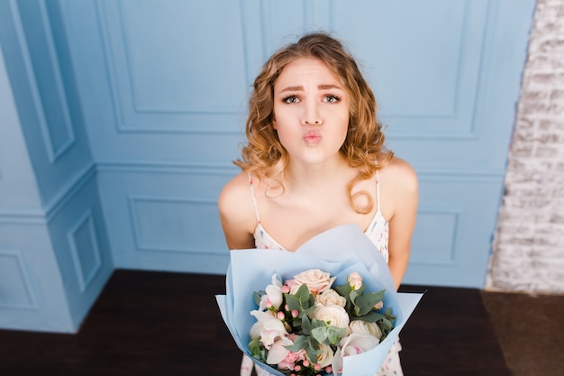 Linda tierna chica rubia de pie en un estudio con paredes azules y sosteniendo un ramo de flores en sus manos