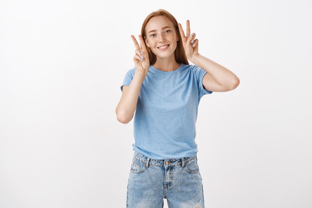 Linda y tierna mujer pelirroja femenina con camiseta azul que muestra signos de paz o victoria cerca de la cara y sonriendo alegremente posando, haciendo una imagen para la red social para enviar amigos