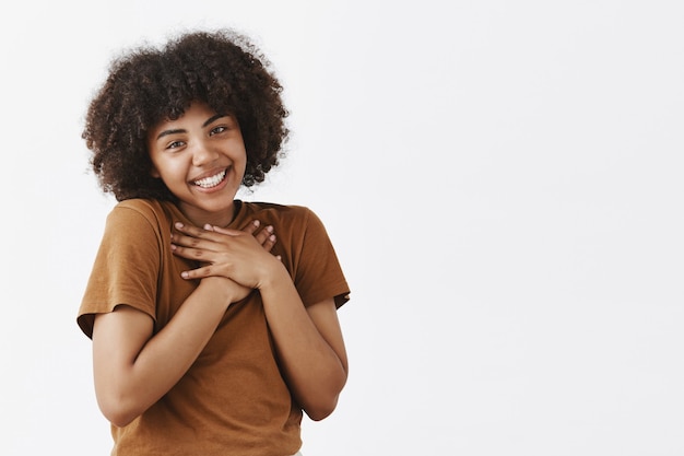 Linda y tierna mujer romántica de piel oscura con peinado afro en camiseta marrón tomados de la mano en el corazón inclinando la cabeza coqueta y sonriente siendo tocada y complacida sobre la pared gris