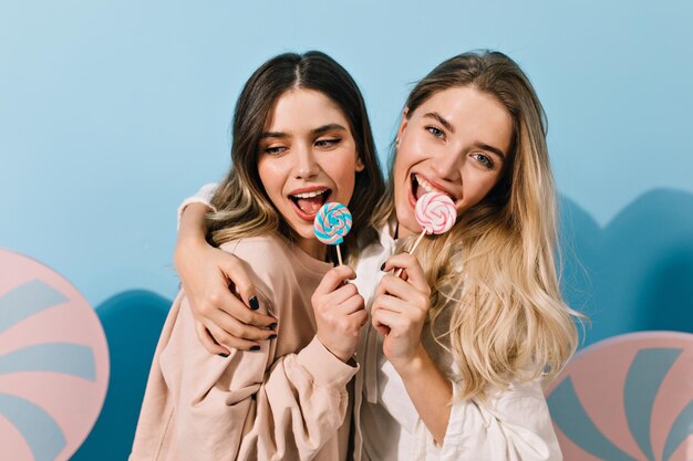 Lindas chicas con dulces abrazando sobre fondo azul Foto de estudio de encantadoras mujeres europeas con piruletas