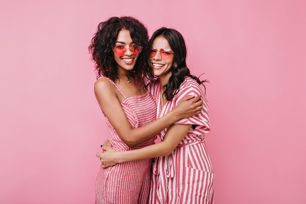 Lindas chicas románticas se abrazan amistosamente. Señoras con gafas de sol rosas riendo.