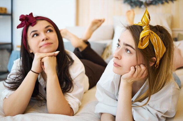 Lindas hermosas chicas adolescentes con cintas para la cabeza acostado en el sofá, tomados de la mano debajo de la barbilla y mirando con expresiones faciales pensativas, sintiéndose aburrido