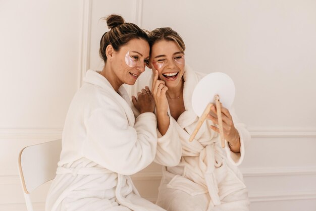 Lindas mujeres jóvenes caucásicas en batas de baño se admiran mientras se miran en el espejo sobre fondo blanco. Belleza y cuidado personal, concepto de cuidado de la piel y la cara