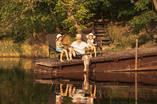 Lindas niñas y su abuelo están pescando en el lago o en el río. Descansando en el muelle cerca del agua y el bosque en el atardecer del día de verano. Concepto de familia, recreación, infancia, naturaleza.