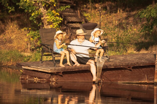 Lindas niñas y su abuelo están pescando en el lago o en el río. Descansando en el muelle cerca del agua y el bosque en el atardecer del día de verano. Concepto de familia, recreación, infancia, naturaleza.