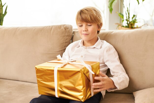 Lindo adolescente de raza caucásica sentado en el sofá con el regalo de año nuevo en su regazo. Chico guapo listo para abrir caja dorada con regalo de Navidad, habiendo curioso anticipado expresión facial, sonriendo