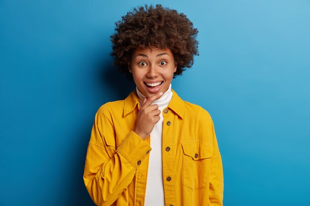 Lindo afroamericano sonriente, mira con expresión de sorpresa y alegría a la cámara, escucha noticias agradables, viste una chaqueta amarilla brillante, aislada sobre fondo azul.