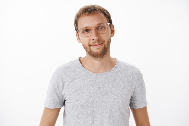 Lindo, amable y guapo, europeo masculino con barba en gafas y camiseta gris sonriendo felizmente con expresión relajada y aliviada escuchando la pregunta del cliente sobre una pared blanca