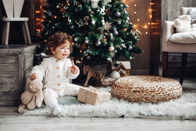 lindo bebé en blanco con el dedo hacia arriba sentado bajo el árbol de Navidad con conejo de peluche.