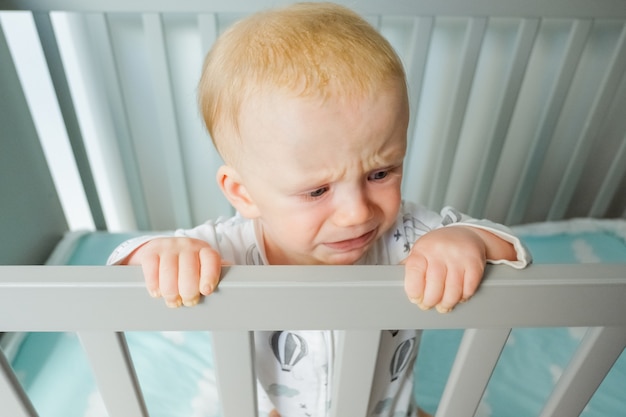 Lindo bebé preocupado de pie en la cuna, sosteniendo la barandilla, llorando y mirando a otro lado. Primer disparo, ángulo alto. Concepto de infancia o cuidado infantil