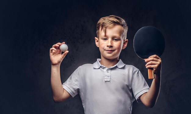 Lindo chico pelirrojo vestido con una camiseta blanca sostiene una raqueta de ping-pong y una pelota en un estudio. Aislado en un fondo de textura oscura.