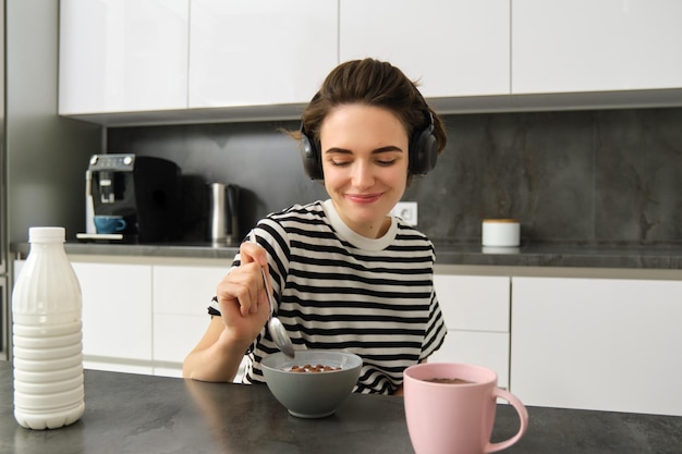 Lindo estudiante mujer moderna comiendo un desayuno rápido teniendo cereales con leche y café escuchando